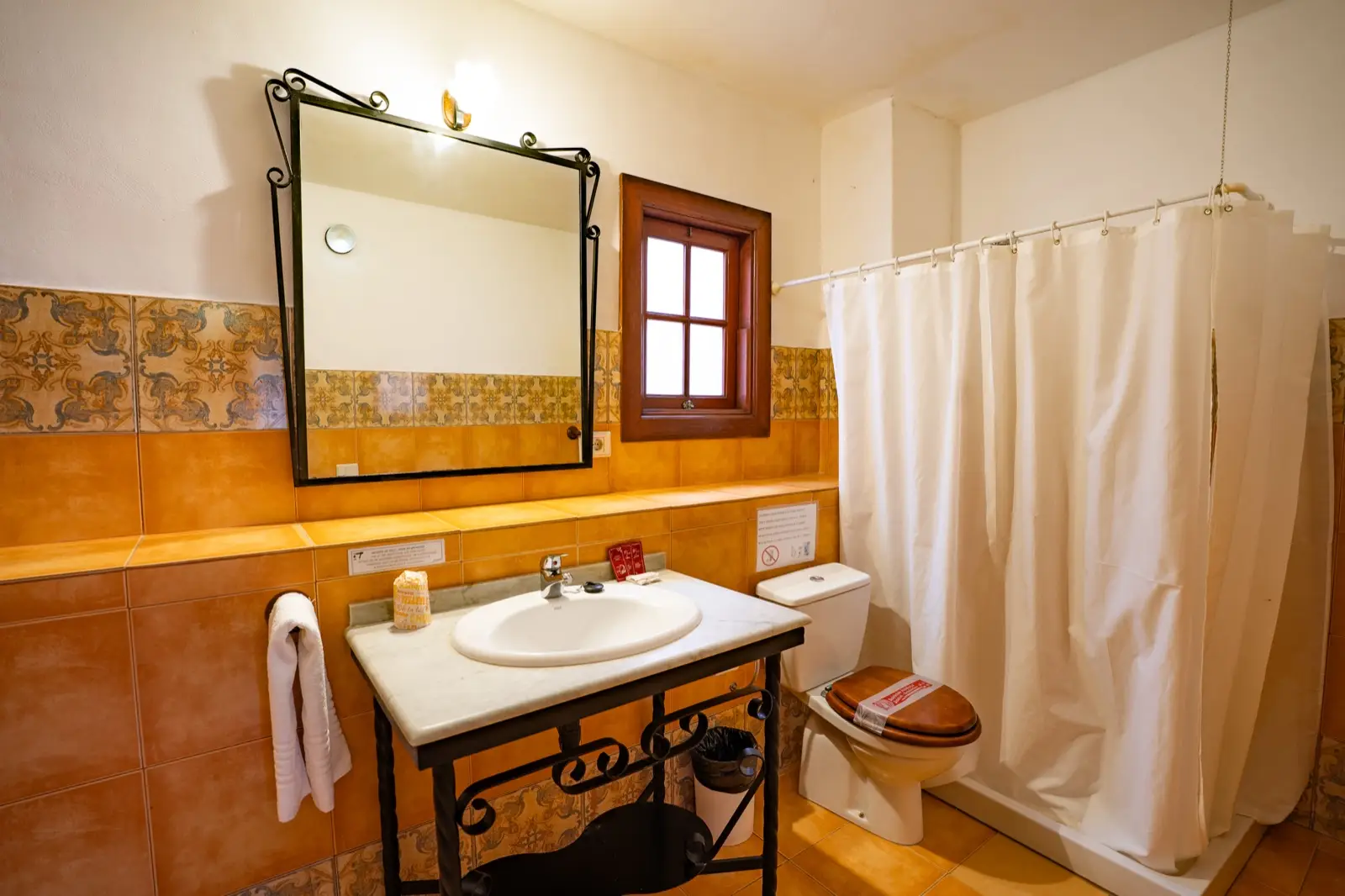 Baño con lavabo de mármol sobre pie de forja, espejo con marco de hierro, ventana de madera, azulejos ocres y ducha con cortina