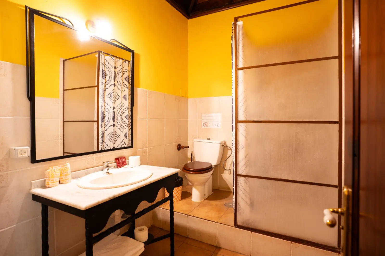 Baño con paredes amarillas, lavabo de mármol sobre pie de forja, espejo con marco de hierro y ducha con mampara de cristal translúcido