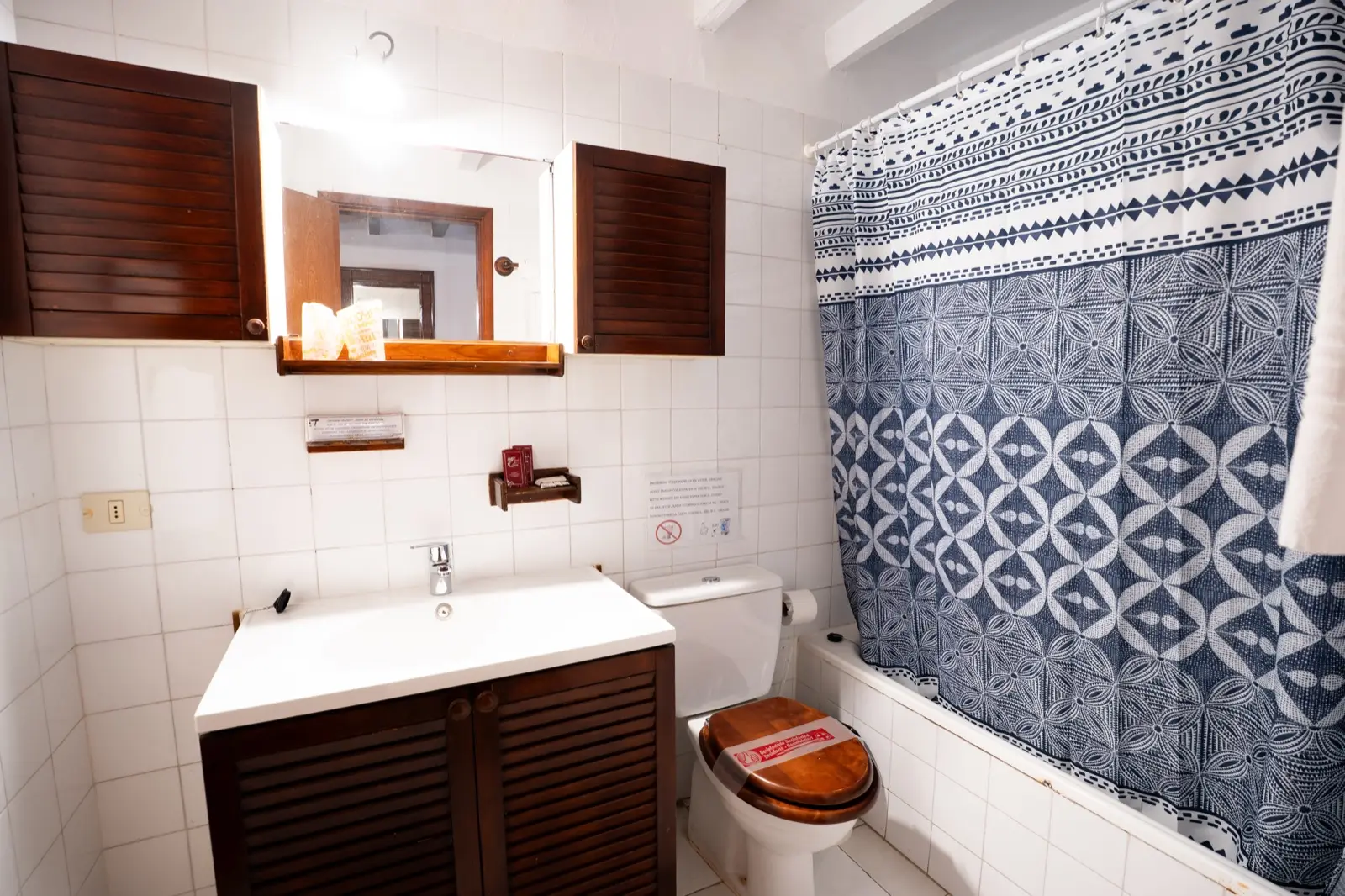 Baño de la habitación triple: azulejos blancos, lavabo sobre mueble de lamas de madera, espejo entre dos armarios de madera y bañera con cortina estampada en azul y blanco
