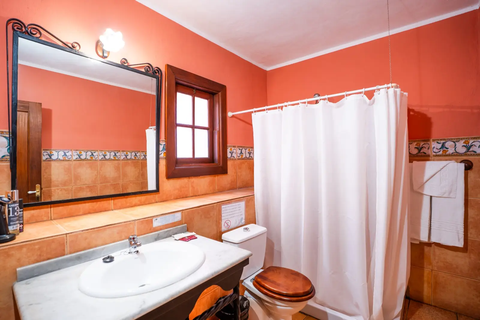 Baño de una habitación doble con cama de matrimonio: paredes color teja, zócalo de azulejos con cenefa, lavabo de mármol, espejo con marco de forja, ventana de madera, toallas y cortina blanca