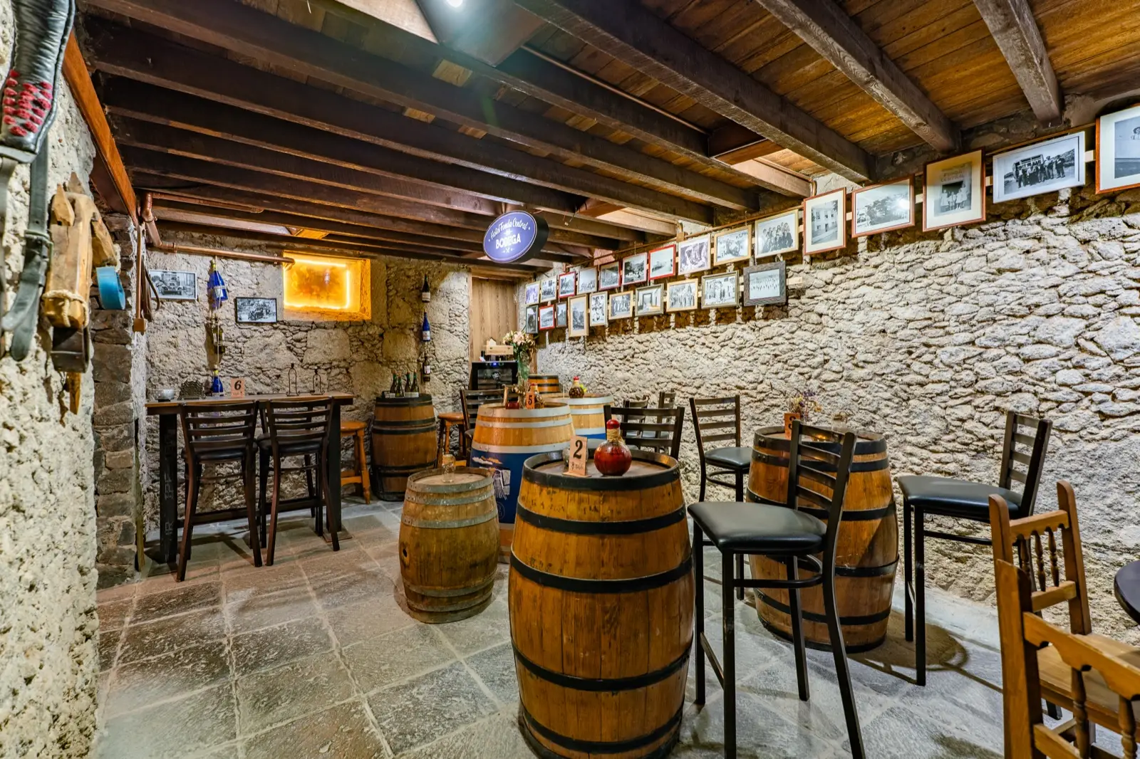 Bodega con barricas como mesas, sillas altas, techo de vigas de madera y una pared de piedra con fotografías antiguas