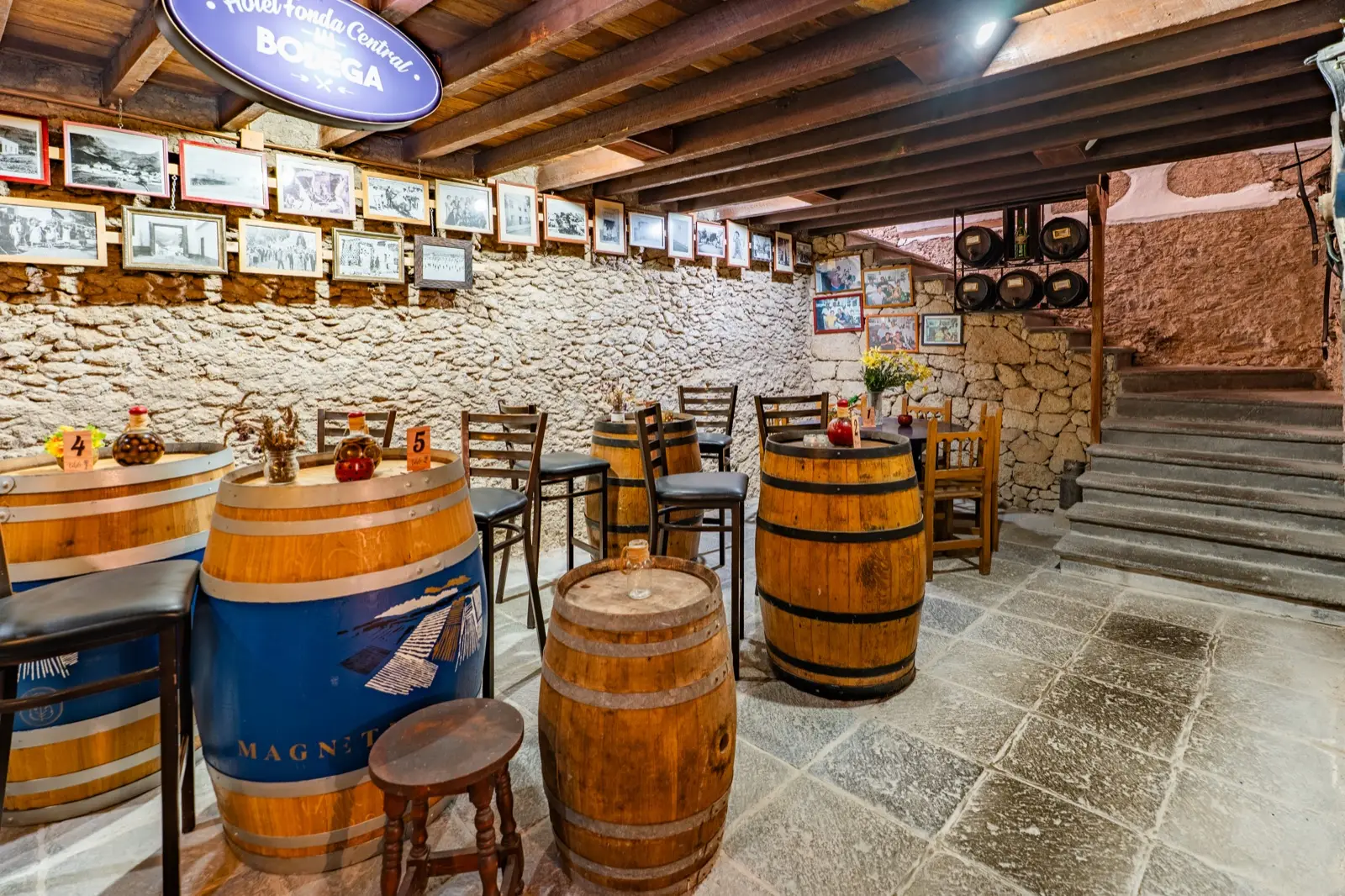 Bodega con paredes de piedra, barricas de madera como mesas, taburetes altos, fotografías antiguas enmarcadas y el cartel «Hotel Fonda Central Bodega»