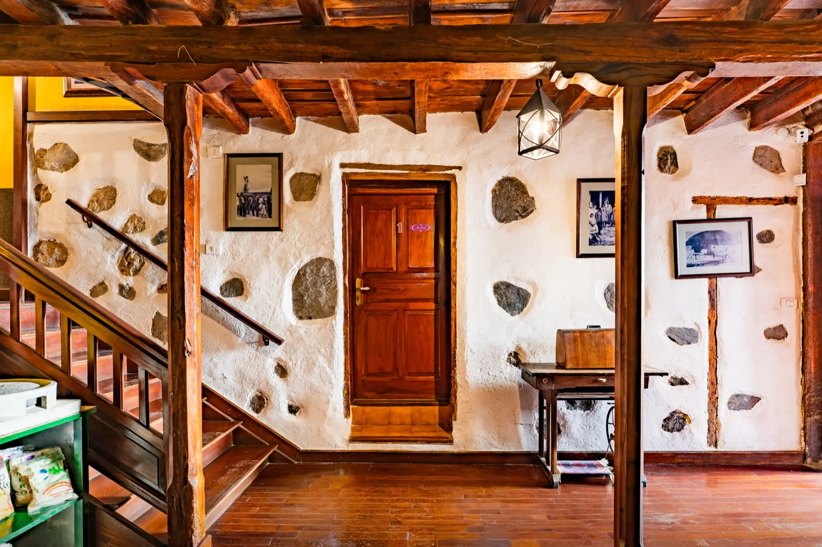 Zona de paso con paredes encaladas de piedra vista, pies derechos y vigas de madera, escalera de madera y la puerta de la habitación 1