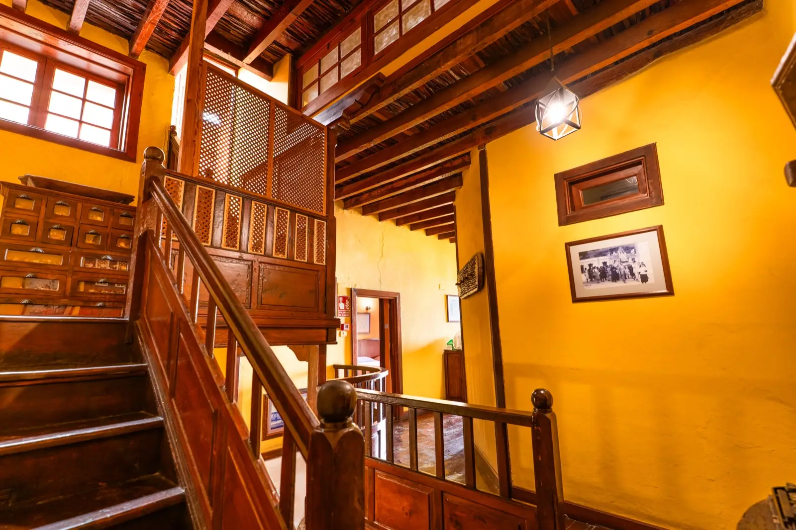 Escalera de madera con celosías, paredes amarillas, techo de vigas y fotografías antiguas enmarcadas