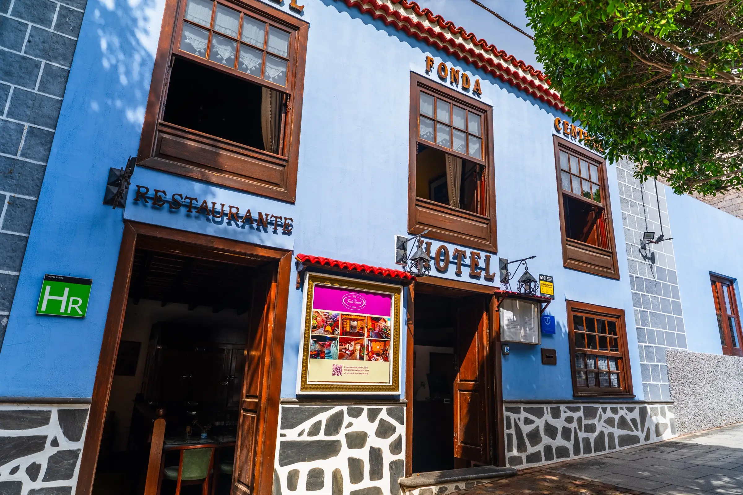 Fachada azul del Hotel Fonda Central en la calle Grande de Adeje, con ventanas de madera, zócalo de piedra y las entradas del restaurante y del hotel