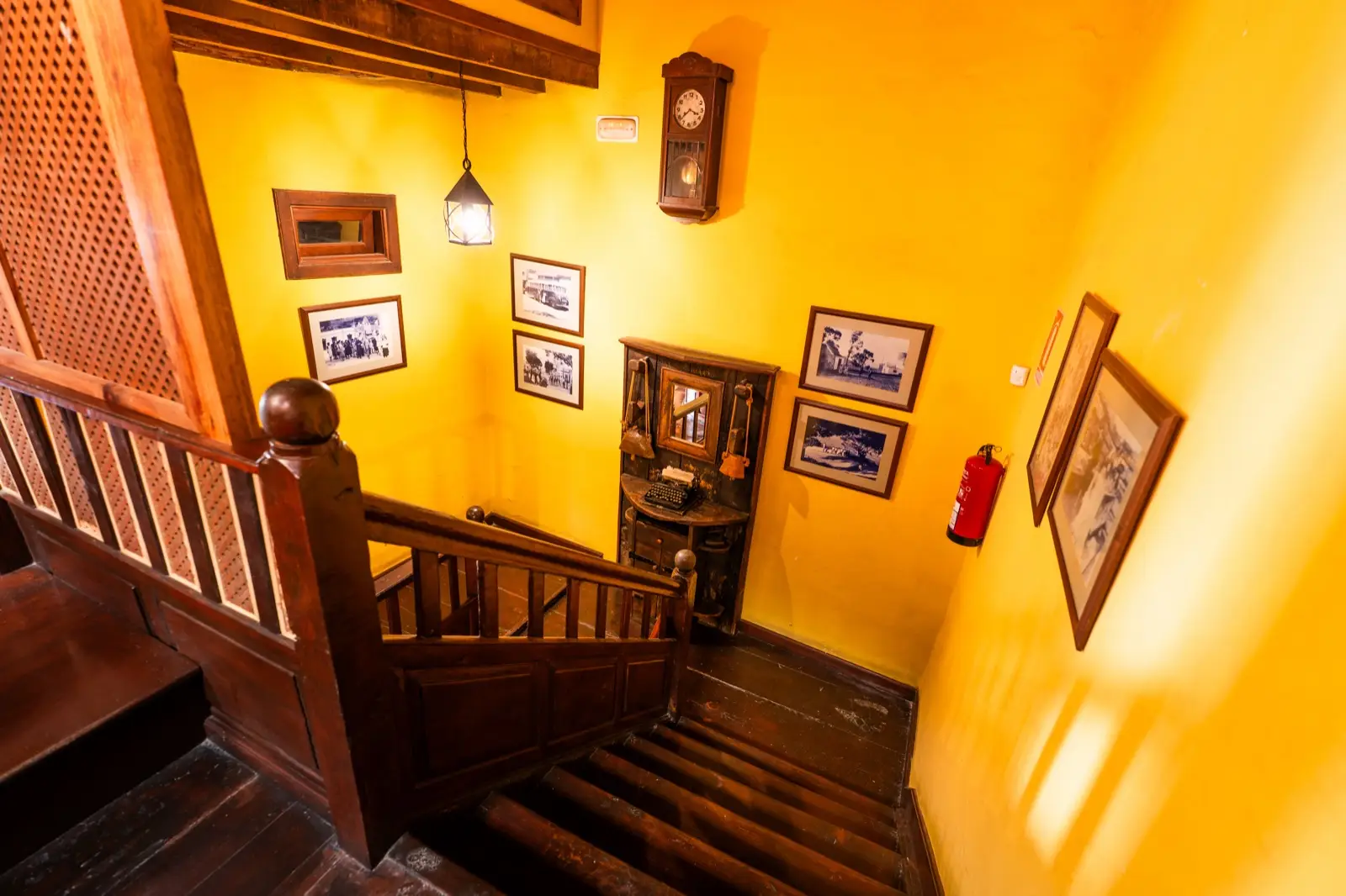 Escalera de madera vista desde arriba, con paredes amarillas, un reloj de pared y fotografías antiguas enmarcadas