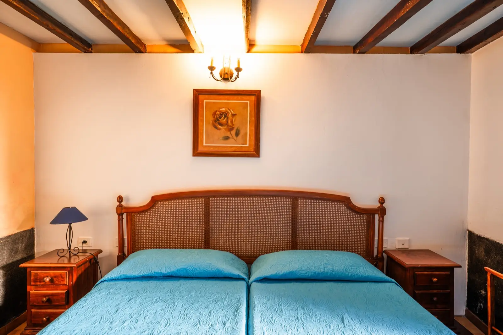 Habitación triple: dos camas individuales juntas con colcha azul, cabecero de rejilla, mesitas de madera y techo de vigas