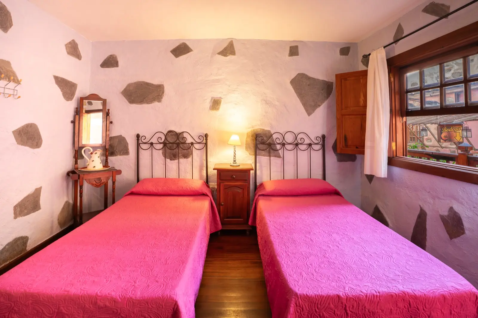 Habitación doble con dos camas individuales, cabeceros de forja, colchas fucsia y paredes encaladas con piedra vista