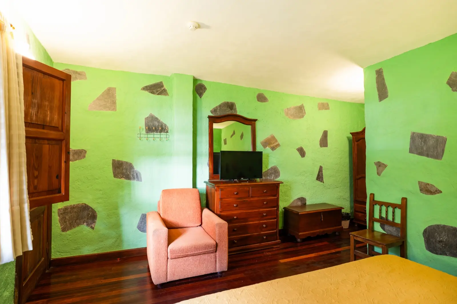 Habitación doble con sillón, cómoda con televisor, baúl de madera y paredes verdes con piedra vista