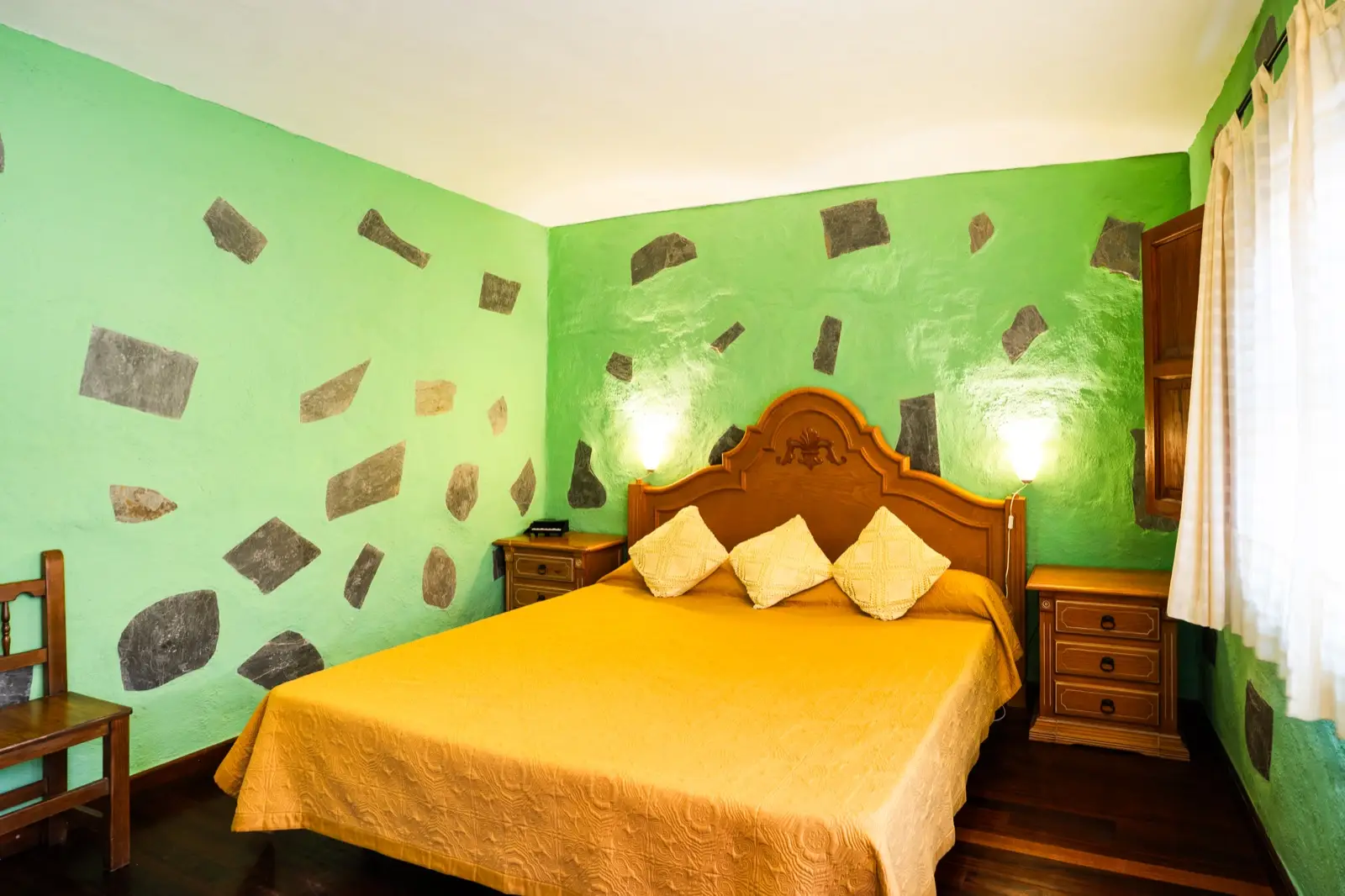 Habitación doble con cama de matrimonio, cabecero de madera tallada, colcha dorada y paredes verdes con piedra vista