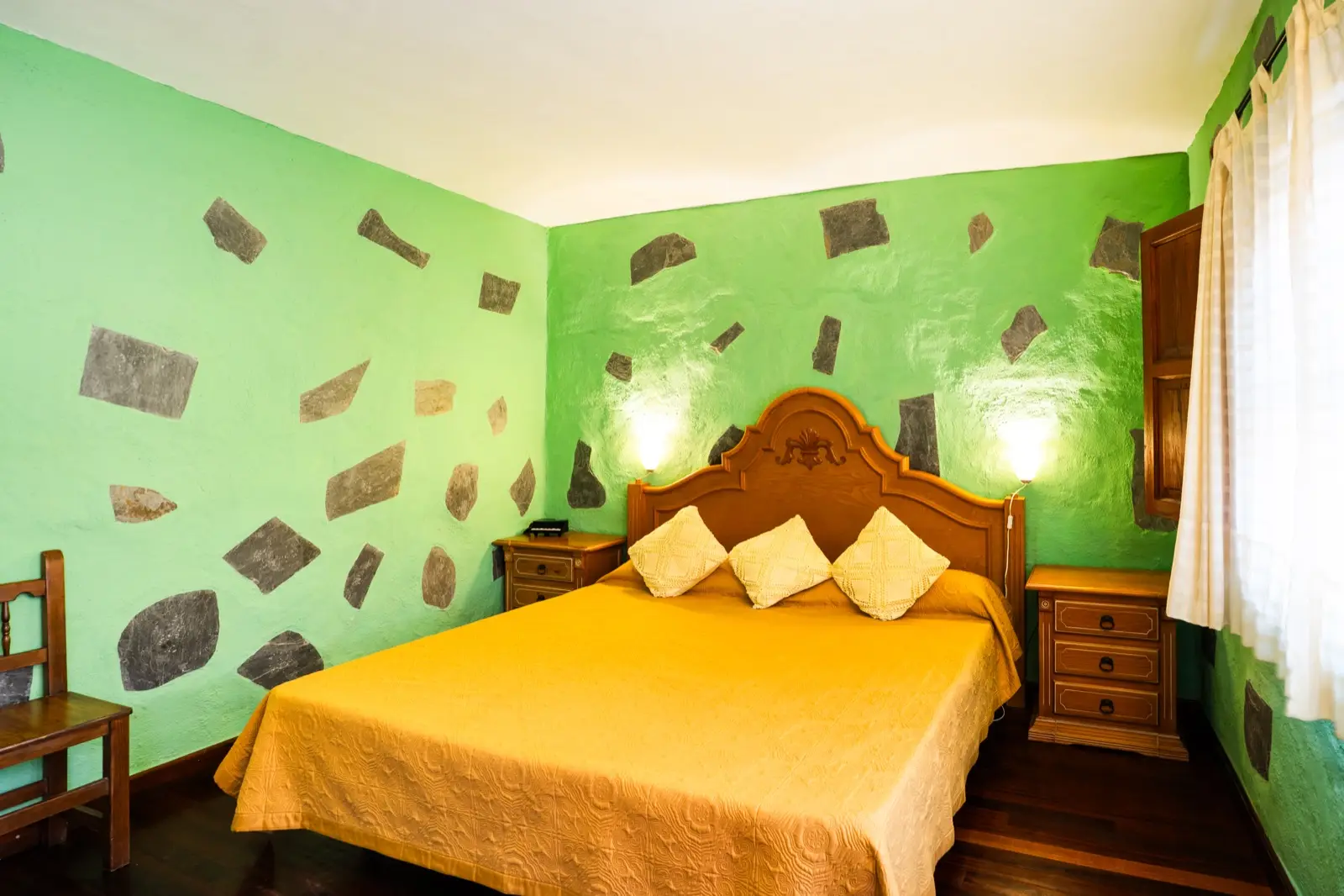 Habitación doble con cama de matrimonio, cabecero de madera tallada, colcha dorada y paredes verdes con piedra vista