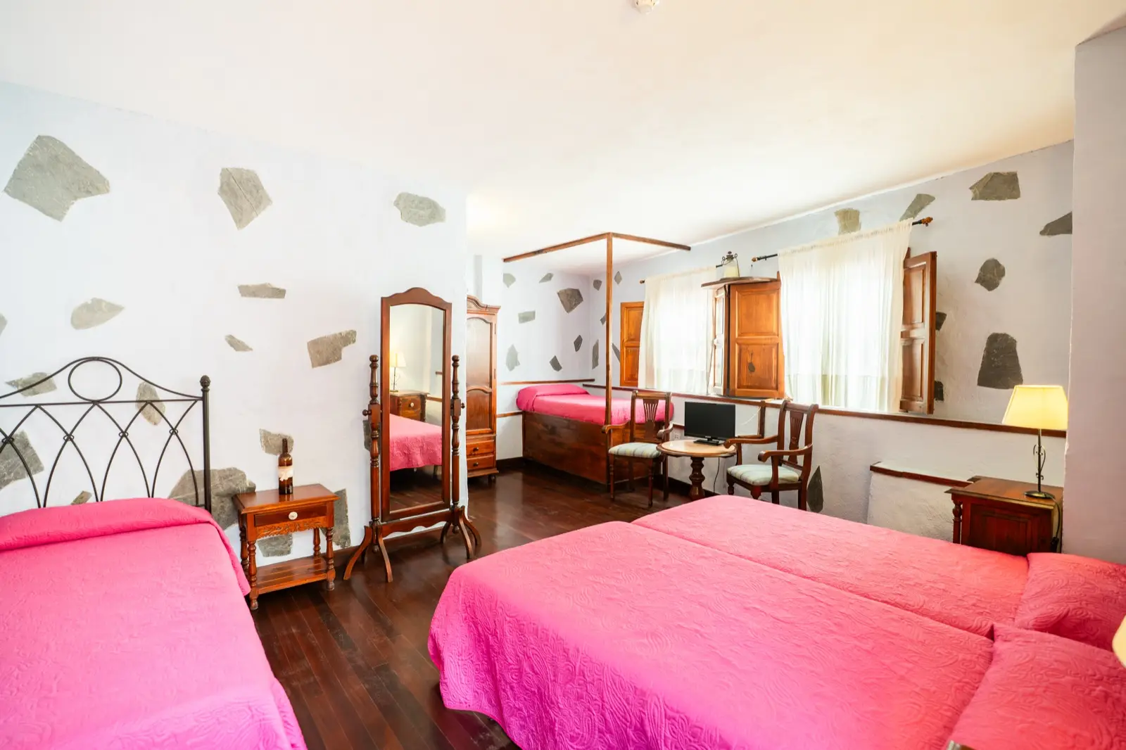 Habitación cuádruple vista de conjunto: camas individuales, espejo de pie, mesa con dos sillas y ventanas con contraventanas de madera