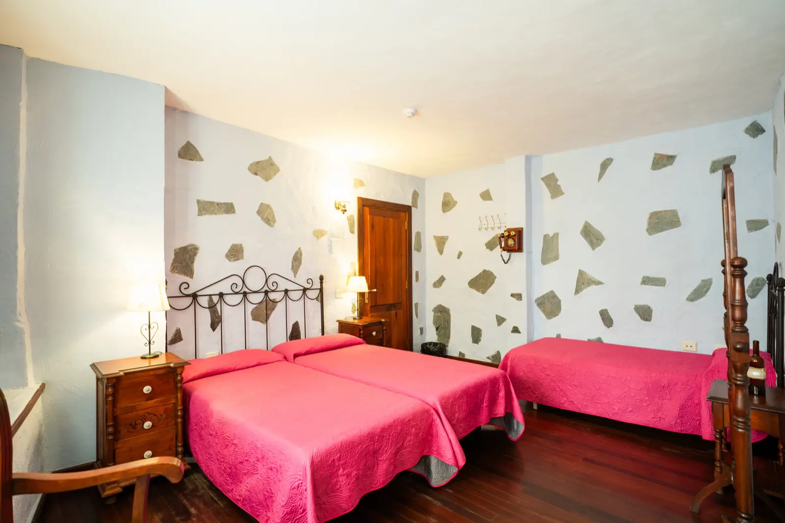 Habitación cuádruple con camas individuales de colcha fucsia, cabeceros de forja y paredes blancas con piedra vista