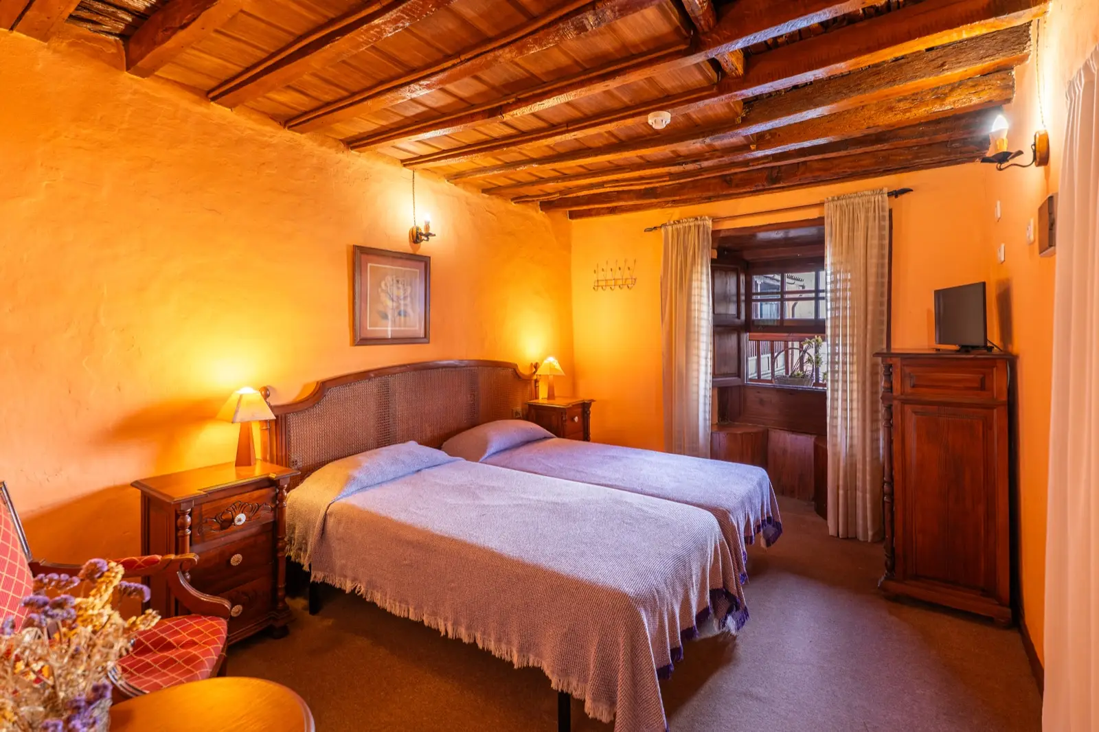 Habitación doble con dos camas individuales juntas de colcha lila, techo de vigas de madera y ventana de madera