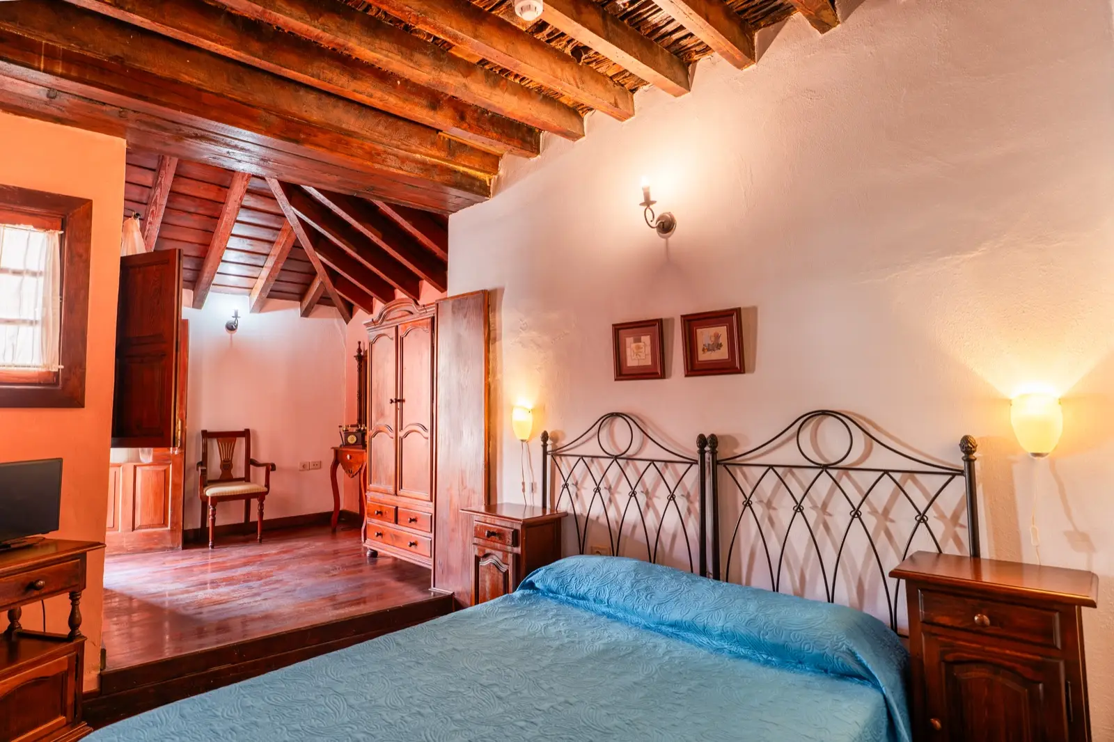Habitación doble con cama de matrimonio de colcha azul, cabecero de forja, techo de vigas de madera y una zona elevada con armario y silla