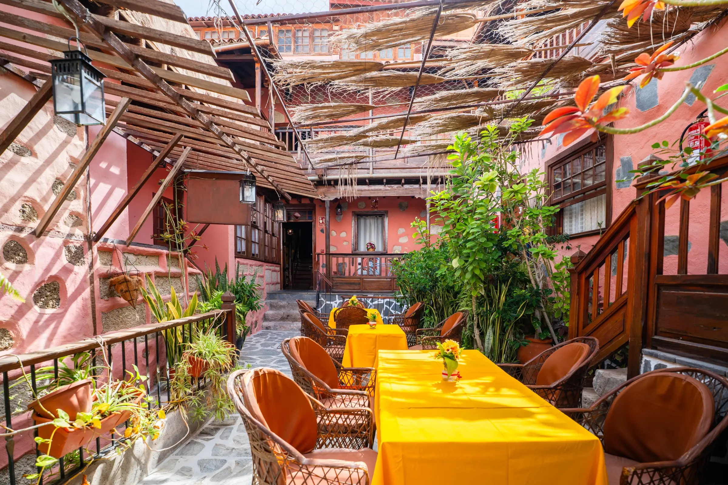 Patio interior de la casona con mesas de mantel amarillo, sillones de mimbre, pérgola de madera y cañizo, plantas y galerías de madera al fondo