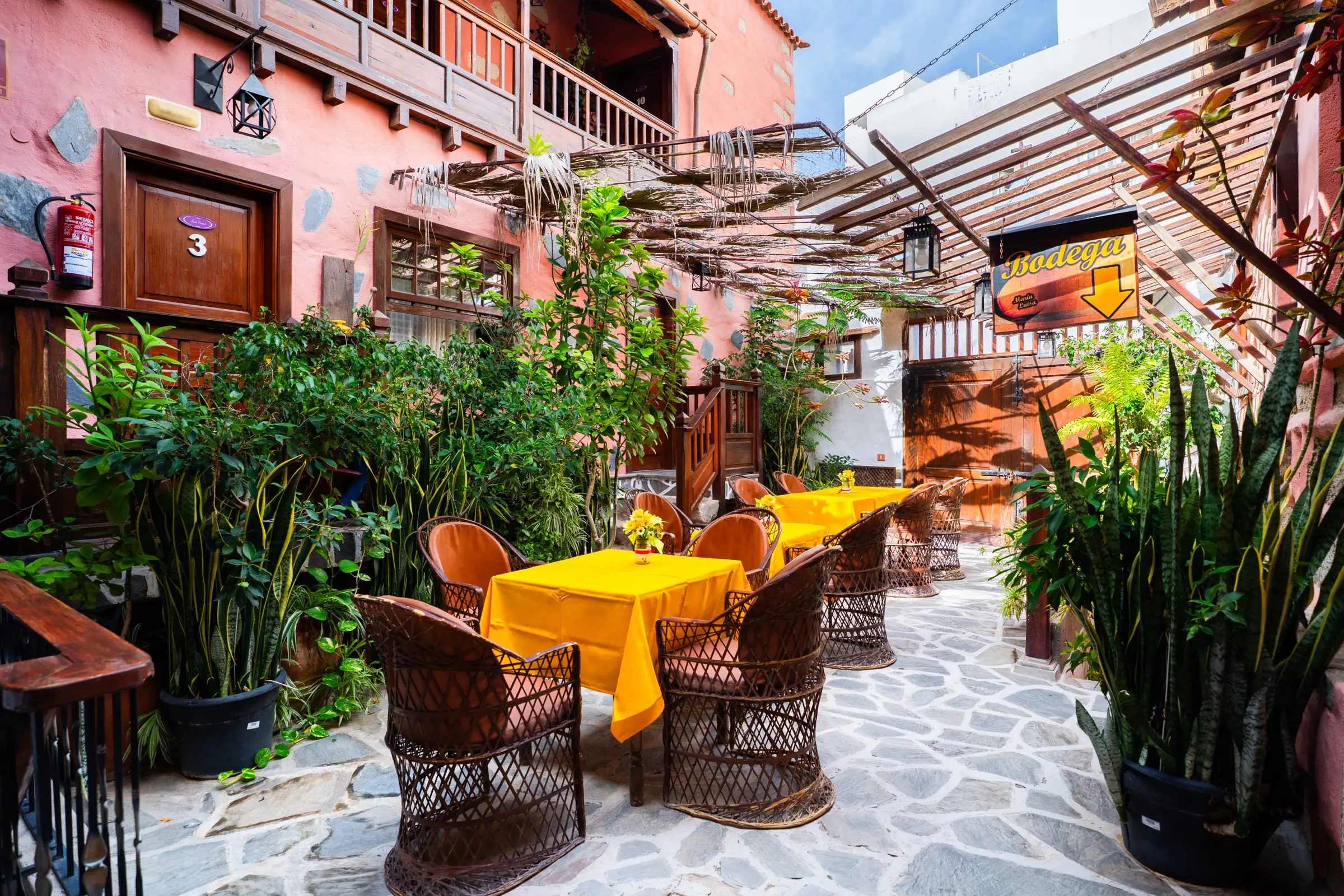 Patio con mesas de mantel amarillo y sillones de mimbre entre plantas, bajo una pérgola de madera, con el cartel de la bodega al fondo